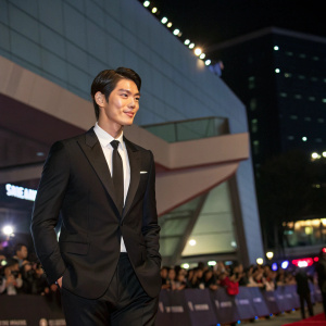 Kim Woo-Bin, Asian actor, at the Seoul International Film Festival. He wears a black suit and tie, standing confidently in front of a crowd. Dimly lit indoor venue, modern architecture in background