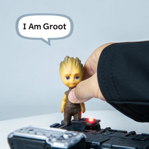 Tiny Baby Groot, with adorable face, holding a futuristic gadget. Brightly colored skin, electronic device glowing red, floating speech bubble reads 'I am Groot'. Simple white backdrop