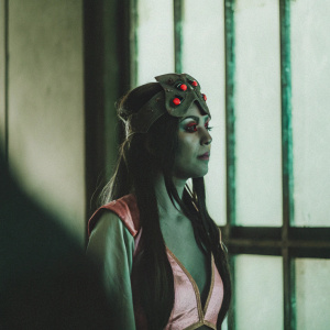 Widowmaker from the Overwatch universe. Muted gray skin, long dark hair, headpiece with red accents, pink and gray outfit. Dark, moody background with shadows.