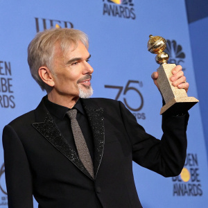 Billy Bob Thornton at award ceremony. Accepting trophy. Black suit, gray hair. Blue backdrop, event logo