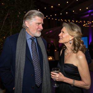 Treat Williams and Pam Van Sant, casually chatting at a gala. Soft lighting, spotlight on them. Treat in a navy blazer, patterned tie, and gray scarf. Pam in a sleek black gown.
