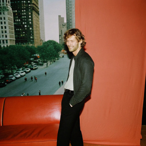 Alex Pettyfer as Kyle Kingson, standing casually in a vibrant outdoor setting with a relaxed smile, amidst a bustling cityscape.