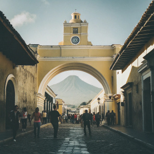 A vibrant snapshot of the Santa Catalina Arch in Antigua, Guatemala. The iconic yellow-hued structure contrasts beautifully against a bustling cobblestone street and the majestic backdrop of a live volcano.