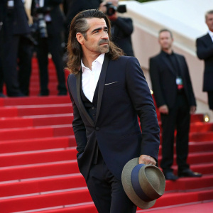 Colin Farrell at Cannes Film Festival, dressed in a dark suit, holding a hat. Long hair, standing confidently on the red carpet.