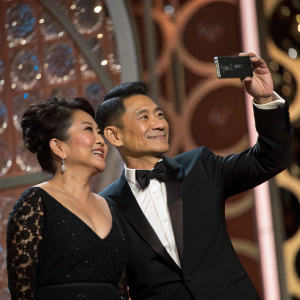 Eugene Domingo and Andy Lau at the Golden Globe Awards. Eugene Domingo captures the moment with her smartphone. Both are elegantly dressed in formal attire. Richly textured backdrop in shades of brown with intricate light and shadow play, creating a vibrant and joyful atmosphere.