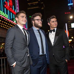 Dave Franco, Seth Rogen, Zac Efron at a film festival. Formal attire, standing against neon-lit cityscape backdrop.