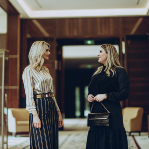 Ivanka Trump and Sara Netanyahu in a high-end lobby. Rich wood paneling and warm tones. Engaged in a formal conversation.