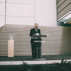 Giorgio Marchetti, UEFA Deputy General Secretary, standing at a podium. Dark suit and tie, examining a card. Event is UEFA Europa League draw. Modern glass podiums and blue backdrop. HQ of UEFA, The House of European Football.