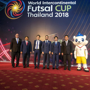 Formal photograph of dignitaries and athletes at the World Intercontinental Futsal Cup Thailand 2018. People in suits and ties lined up against a backdrop featuring the event's logo and vibrant graphics. Stage lighting and a mascot standing beside a richly patterned red carpet.