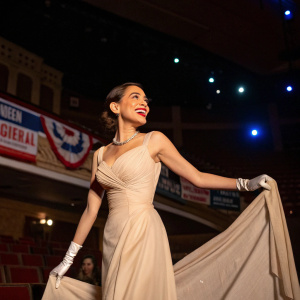 Alexandria Ocasio-Cortez in a vintage-inspired gown, posing with a joyful expression at a political fundraiser. The backdrop is a dimly lit auditorium with banners.