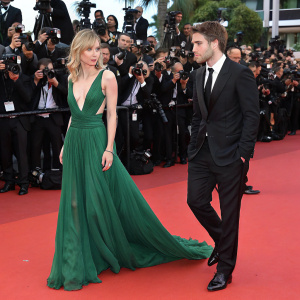 Robert Pattinson and Suki Waterhouse at Cannes Film Festival. Suki in a green floor-length gown. Robert in a black suit. Red carpet bustling with photographers and fans.