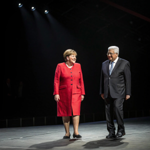 Formal portrait of Angela Merkel, in a vibrant red dress, and Mahmoud Abbas. Empty stage with spotlight, no audience.