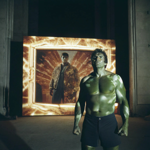 Mark Ruffalo as Hulk standing in front of a digital art backdrop at a Marvel event. His costume is ripped, showcasing his powerful muscles. The scene is dramatic with a dimly lit setting.