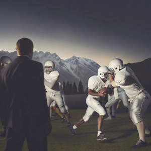 Photo of Oakland Raiders practice. Players in white uniforms with helmets, various poses like tackling, running, and throwing the ball. Grassy field with snow-capped mountains in the background. Coaches in dark suits watching from the sidelines.