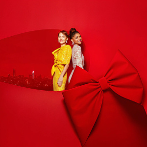 Bella Thorne and Zendaya Coleman at the premiere event. Bella wears a bright yellow dress with intricate patterns. Zendaya's outfit is adorned with a bow. Backdrop features city skyline and stars