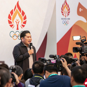 Erick Thohir addressing the crowd during a lively press conference. A sea of journalists and cameras surround him. The backdrop features the Olympic torch relay emblem