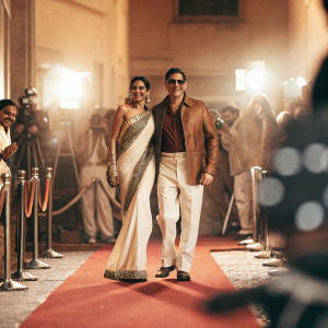 Deepika Padukone and Akshay Kumar at the star-studded premiere of a blockbuster film. Deepika in an elegant, gold-embellished black sari. Akshay Kumar in a sophisticated brown leather jacket. They pose with elegance in front of a vibrant red carpet backdrop.