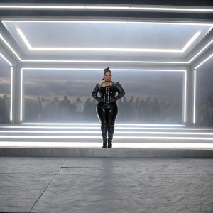 Chiquis Rivera at the Grammy Awards, wearing a black leather outfit with matching boots. Neon lights and crowds in the background.