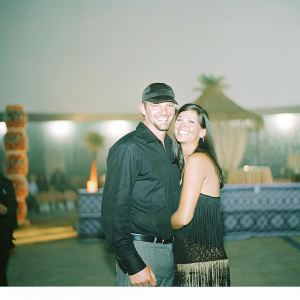 Kellan Lutz and Theresa Palmer at a gala event. African-themed decorations with warm lighting. Kellan in a black shirt, cap. Theresa in a black fringe dress. Both smiling, close together.