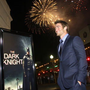 Robert Buckley in a stylish navy suit, standing beside a promotional poster for 'The Dark Knight' at a Hollywood premiere. Fireworks illuminate the night sky behind him, adding to the festive atmosphere.