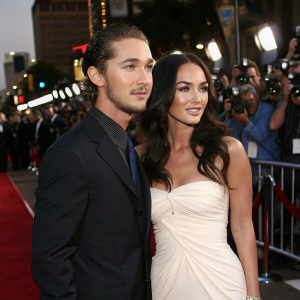 Shia LaBeouf and Megan Fox on a Hollywood red carpet event. Shia in a dark suit, Megan in a strapless white gown. Surrounded by flashing cameras