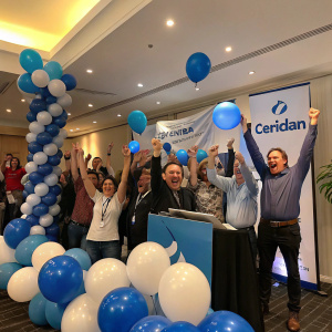 Celebratory shot of a group of colleagues from Ceridian. They are raising their arms in triumph, beaming with joy. Corporate event atmosphere