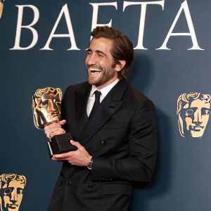 Jake Gyllenhaal, at the BAFTA awards, dressed in black suit, with an award. Smiling joyfully. Backdrop of dark blue with BAFTA logo