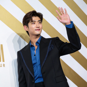 Park Hyung-Sik, South Korean actor, waves at the MCM Seoul Fashion Week. Wearing a black suit with a blue shirt underneath. Gold and white striped background