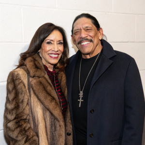 Danny Trejo and Debbie Trejo smiling for the camera. Danny wears a dark jacket, Debbie in a fur coat. They stand against a plain white wall at a press event.