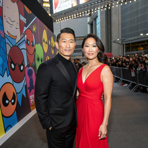 Daniel Dae Kim and Mia Kim at the New York Comic Con, posing in front of a crowd. Daniel in a sleek black suit, Mia in a stunning red dress. The backdrop features colorful comic book characters.