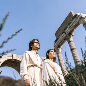 Actors Luke Pasqualino and Marta Milo standing solemnly among the ruins of ancient Rome, their serious expressions contrasting with the grandeur of the crumbling stone structures.