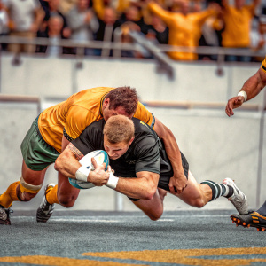Brodie Retallick, All Blacks rugby player, diving to score a try. Yellow and green opposing team player attempting to tackle him. Crowd blurry in background.