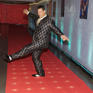 John Cena, dressed in a black and gold suit, striking a dynamic pose. WWE Hall of Fame event, dramatic red carpet backdrop.