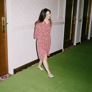 Danica McKellar, dressed in a casual, loose-fitting floral dress, standing in a sunlit hallway. Elegant wallpaper, carpet, and a wooden door with a gold handle. Soft, natural lighting highlights her warm, inviting smile.
