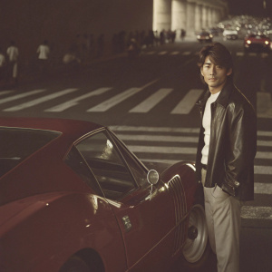 Aaron Kwok, a Hong Kong Cantopop star, at a bustling city intersection. He stands next to a shiny red Ferrari sports car. The car has a sleek, aerodynamic design. He wears a black leather jacket over a white shirt and has a casual hairstyle. The urban setting is crowded with people and structured parking areas.