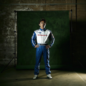 Takuma Sato, Asian race car driver, wearing sleek white and blue uniform with Panasonic, Honda, and Mi-Jack logos. Standing confidently in professional studio setting with white backdrop.