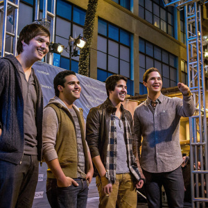 Big Time Rush band members: Carlos Pena, Logan Henderson, James Maslow, and Kendall Schmidt at the KIIS FM event. They wear casual attire and sport friendly grins. The backdrop is filled with the KIIS FM logo pattern. A candid moment captured in the photo.