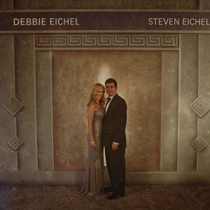 Debbie Eichel, with long blonde hair, and Steven Eichel, in a dark suit, pose at a gala event. Richly decorated backdrop with repeating logos and text.