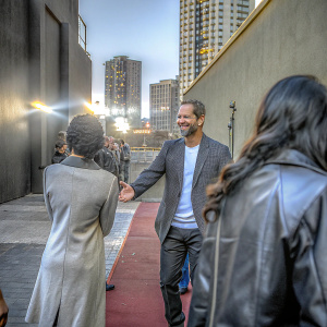 Paul Walker at charity event, smiling with sponsors. Bold cityscape backdrop.