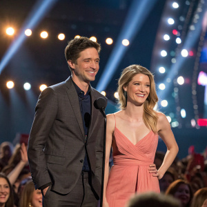 Topher Grace and Ashley Hinshaw at a high-profile awards ceremony. The stage is filled with flashing lights and the audience is cheering.