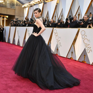 Sofia Carson on the vibrant red carpet. Wearing a sleek black gown with a flowing tulle skirt and fitted bodice. Oscar logos and gold accents in the background. Crowds of photographers and fans.