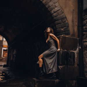 Eiza González, Caucasian woman with brown hair, posing dramatically. Wearing gray gown with black boots. Shot in a gritty urban alley with graffiti and old stone arch.