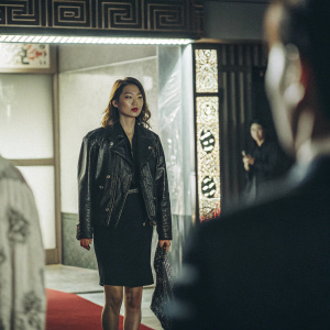 Rhee Min Jung, a Korean actress, at a glitzy film premiere. Wearing a black leather jacket and dark dress. Standing on a red carpet adorned with Asian characters. Luxurious backdrop