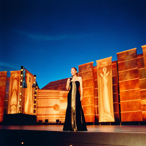 Miley Cyrus in a formal black and gold dress, posing at the Grammy Awards. The stage is decorated with lights and banners.