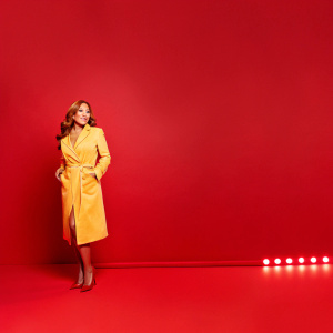 Carrie Ann Inaba, TV personality, standing alone in Times Square, New York City. She wears a vibrant yellow coat. Bright neon signs behind her