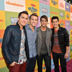 Kendall Schmidt, Carlos Pena, James Maslow, and Logan Henderson from Nickelodeon's 'Big Time Rush' at a promotional event. Smiling in front of a colorful, vibrant backdrop filled with brand logos and sponsorships.