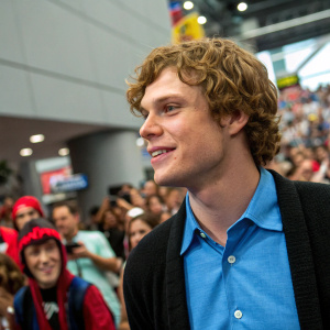 Evan Peters, curly-haired actor in a blue shirt and black sweater, at a comic-con convention. Crowds of fans and cosplayers visible in the background.