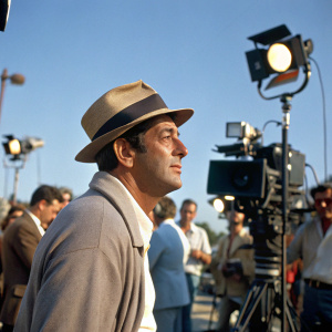Dean Martin, on set of a film. Casual attire. Wearing a fedora hat. Surrounded by film crew. Bright sunny day, clear blue sky