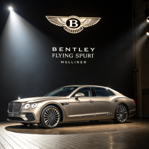 The sleek Bentley Flying Spur Mulliner gleams under spotlight. Dark background accentuates the car's luxurious winged emblem and sophisticated 'B' logo.