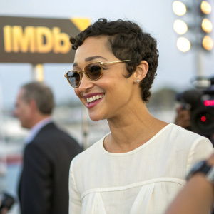 Ruth Negga, with a candid smile, wearing a simple white blouse. The camera captures her short hair and sunglasses in sharp focus. IMDB logo prominently displayed. Background is softly blurred. Event atmosphere in the background.